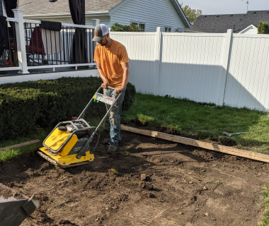 Worker operating a compactor on dirt next to a white vinyl fence