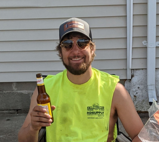Man in neon yellow tank top and ball cap smiling while holding a drink
